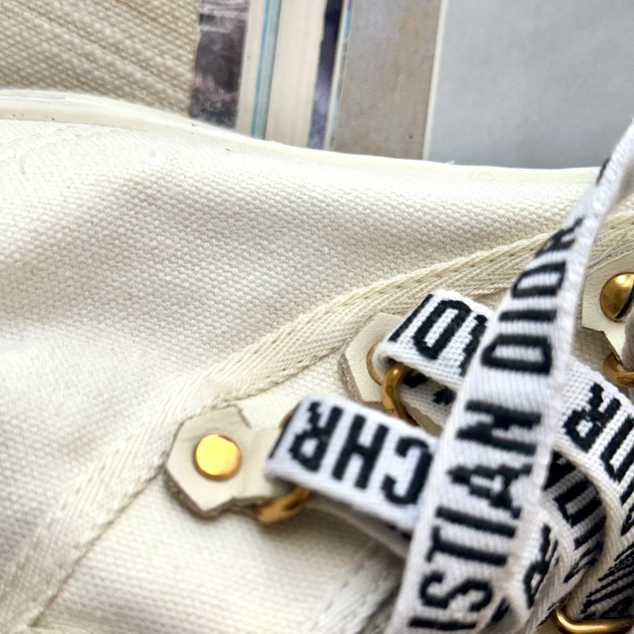 Close-up of a cream canvas bag with gold-tone hardware and a white webbed strap printed with black Christian Dior lettering.