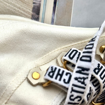 Close-up of a cream canvas bag with gold-tone hardware and a white webbed strap printed with black Christian Dior lettering.