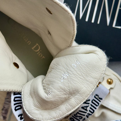 Close-up of a cream-colored leather Christian Dior handbag, showing the interior lining with gold lettering, a white branded ribbon strap, and gold-tone hardware.