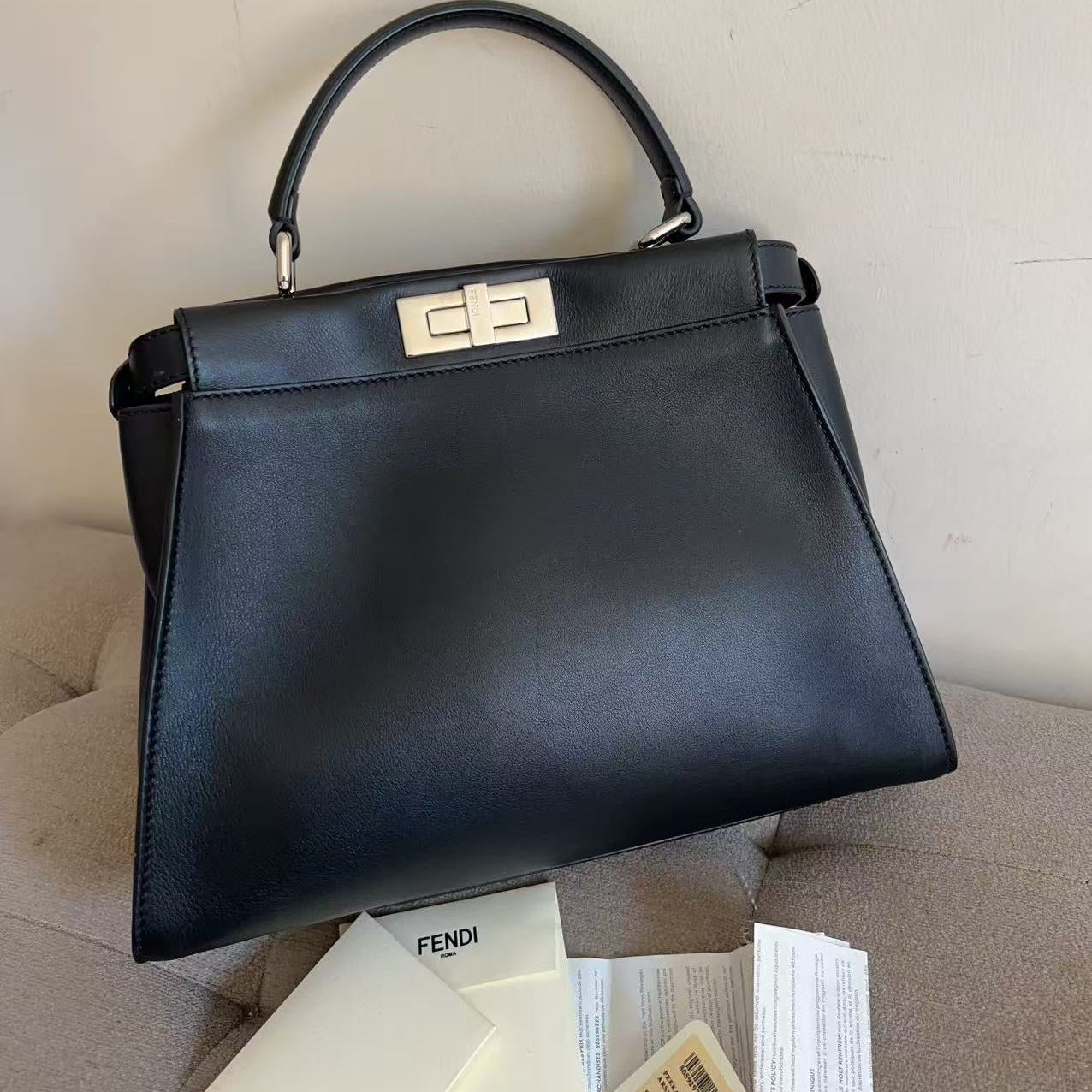 A black leather Fendi handbag with a top handle and gold-tone hardware, placed on a beige upholstered chair with Fendi packaging papers visible below.