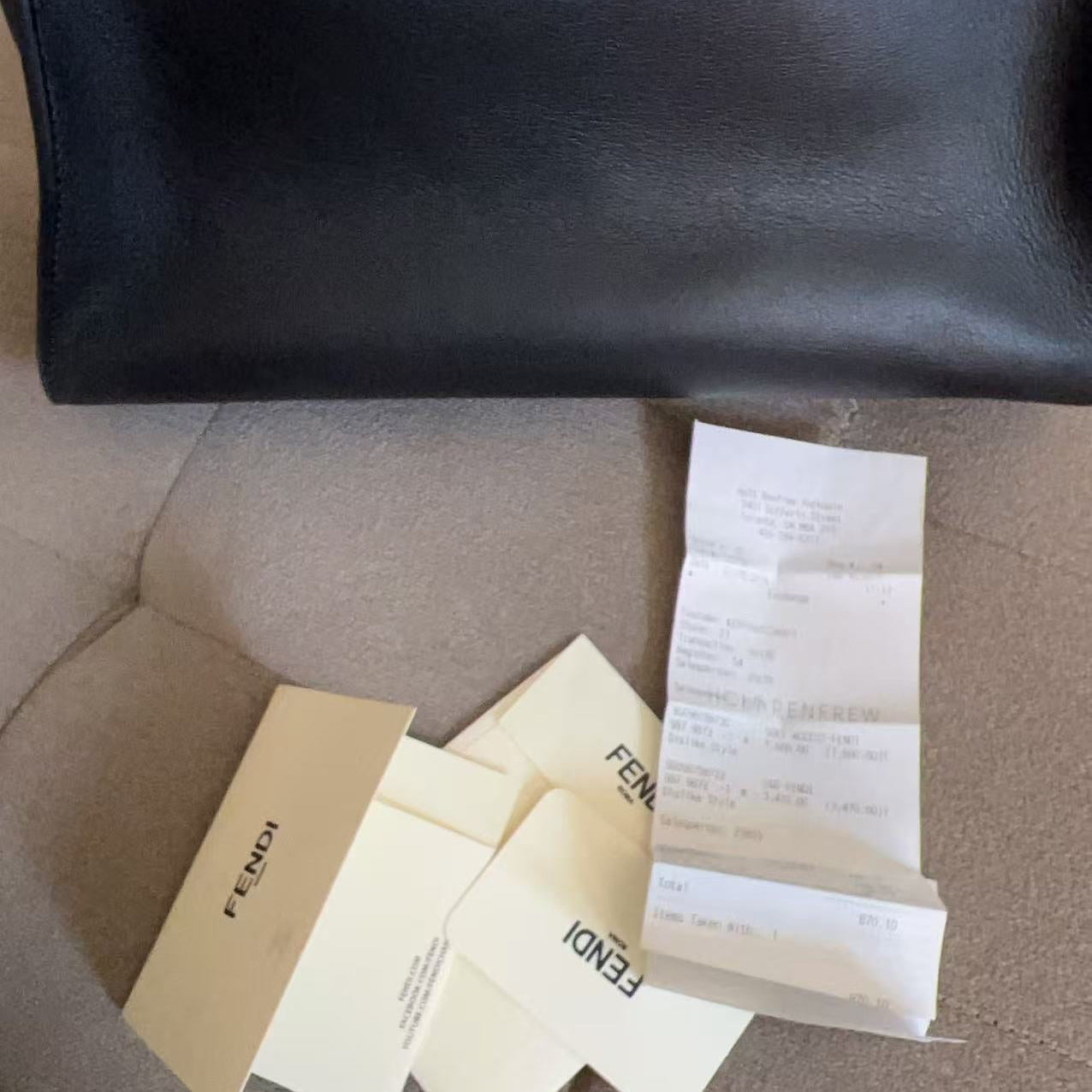 A black leather Fendi handbag is shown with its original packaging and receipt on a gray fabric surface. The packaging includes cream-colored Fendi cards and a receipt.