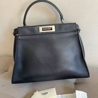 A black leather Fendi handbag with a top handle and a gold-tone turn-lock clasp, resting on a beige upholstered surface with Fendi packaging papers visible in the foreground.