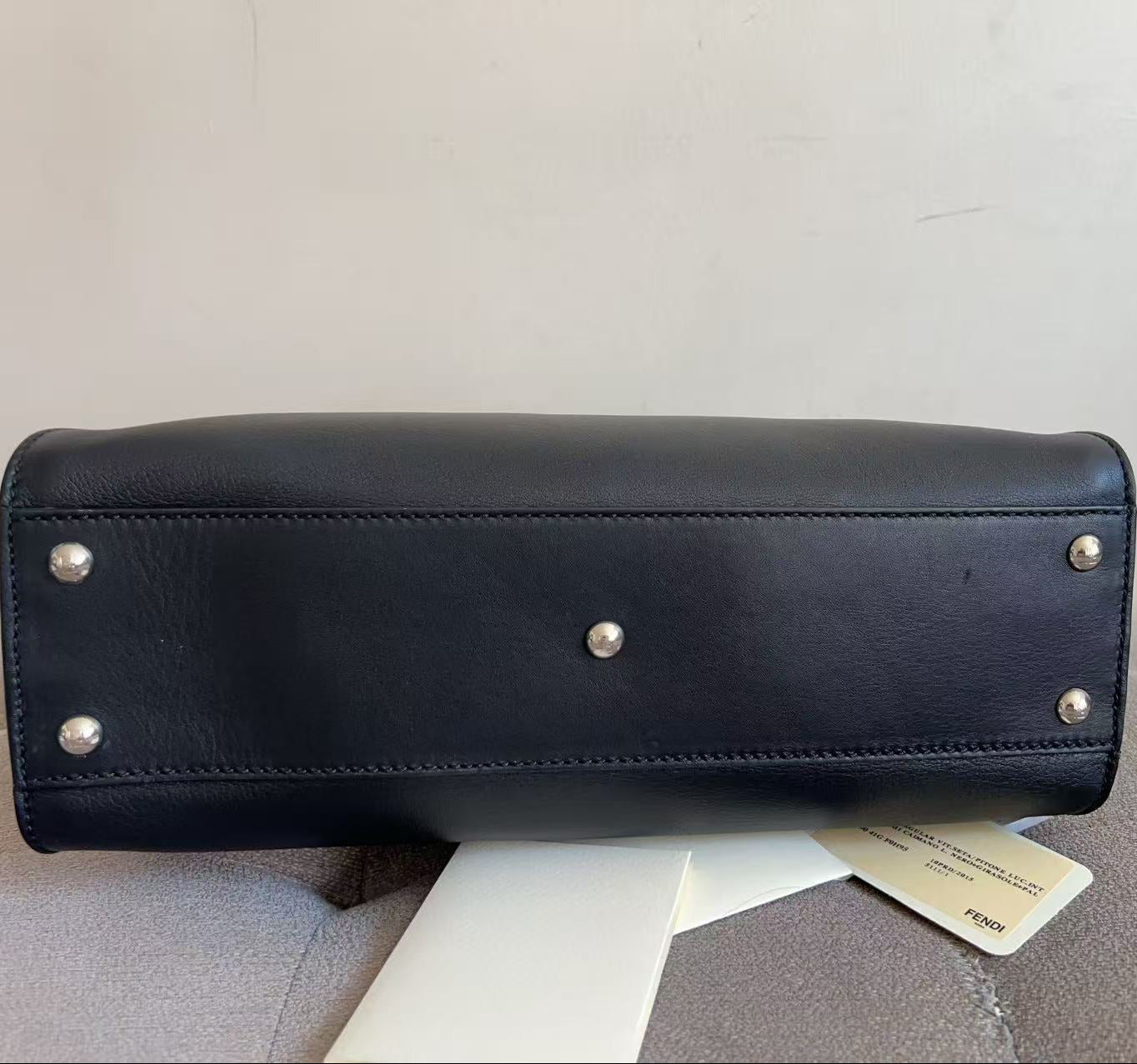 A black leather handbag shown from the bottom, featuring silver-tone metal studs on the base, resting on a light gray upholstered surface with a white tag attached.