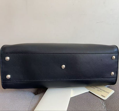 A black leather handbag shown from the bottom, featuring silver-tone metal studs on the base, resting on a light gray upholstered surface with a white tag attached.