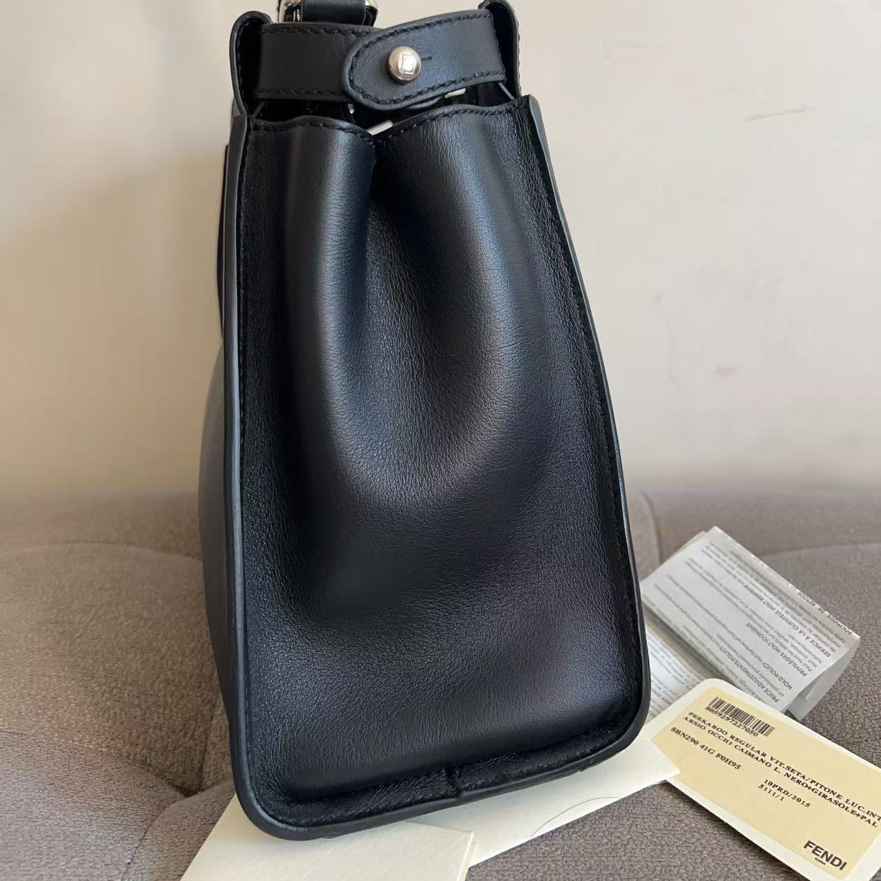 A black leather handbag with a structured top handle and a silver snap button closure, shown standing upright on a light surface with a beige upholstered chair behind it. A small Fendi tag is visible near the base.