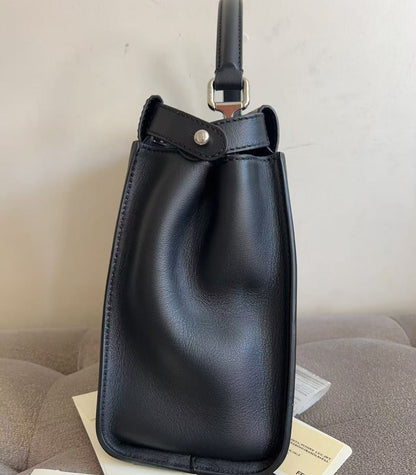 A black leather Fendi bucket bag with a top handle, resting on a gray upholstered chair with its original box and tag visible.
