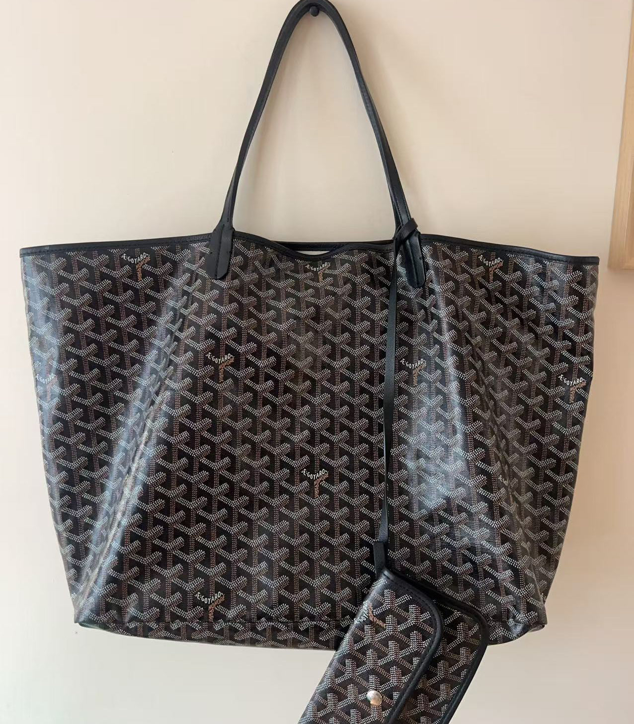 A Goyard black and brown monogram tote bag with matching wallet, hanging against a plain beige wall.