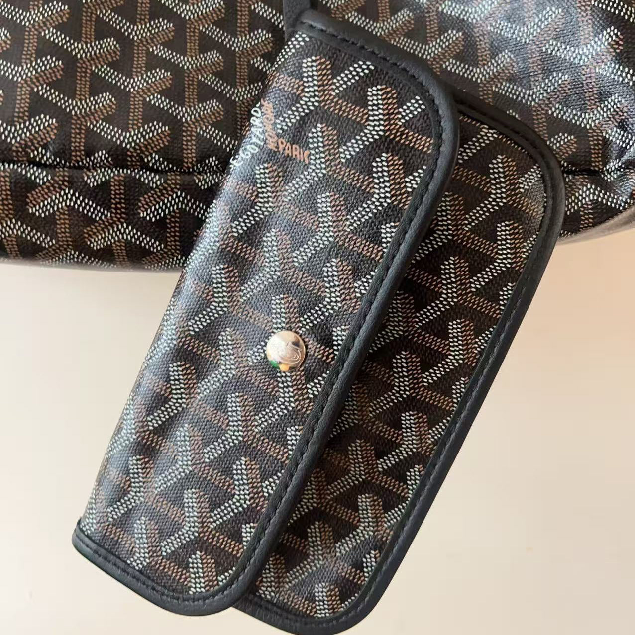 A close-up of a Goyard wallet or pouch in the brand's signature chevron monogram canvas, featuring a black leather trim and snap closure, set against a matching bag.