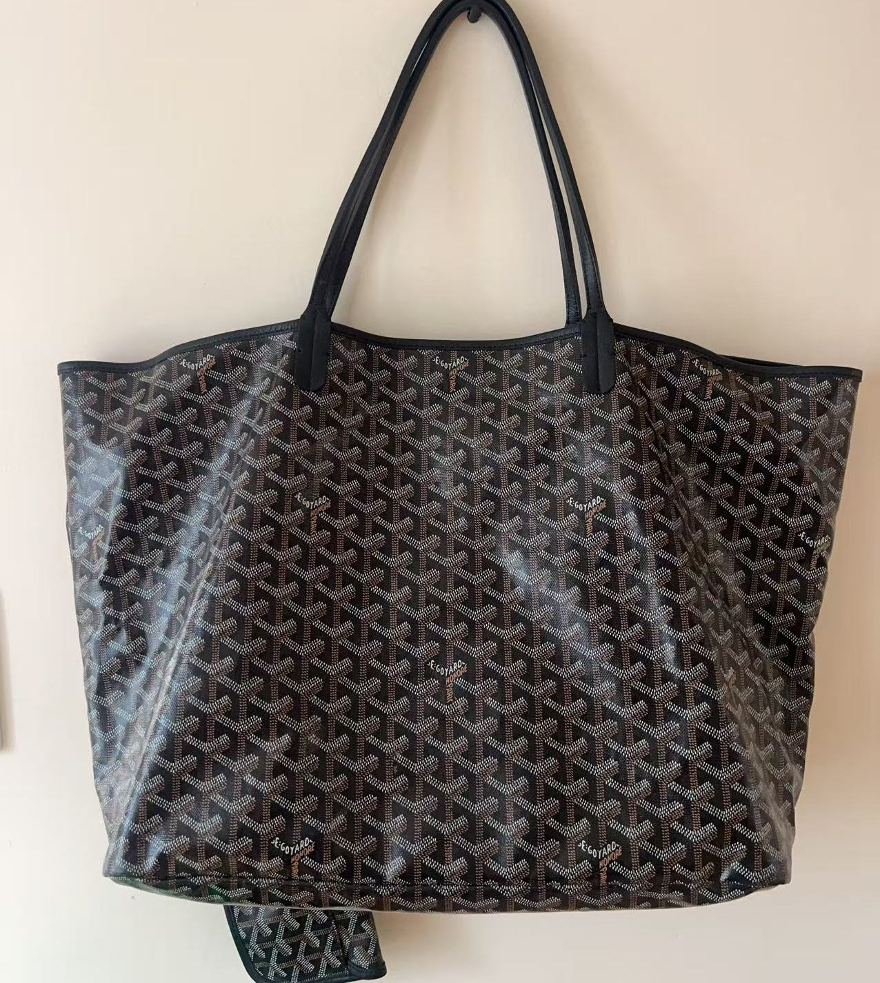 A Goyardine tote bag in black and brown with its signature repeating chevron pattern, featuring black leather handles and a matching zippered pouch hanging from the bottom, set against a plain beige wall.
