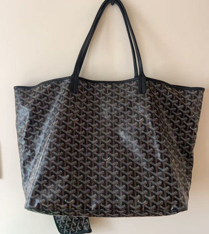 A Goyardine tote bag in black and brown with its signature repeating chevron pattern, featuring black leather handles and a matching zippered pouch hanging from the bottom, set against a plain beige wall.