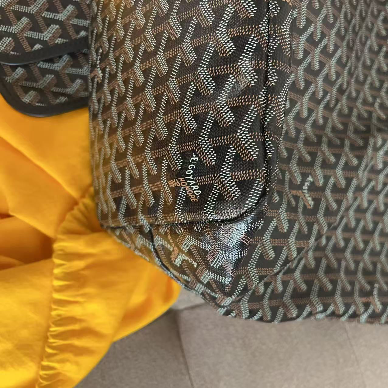 A close-up of a Goyard tote bag featuring the brand's signature chevron monogram pattern in brown, beige, and black. The bag is resting on a light beige sofa next to a bright yellow fabric.