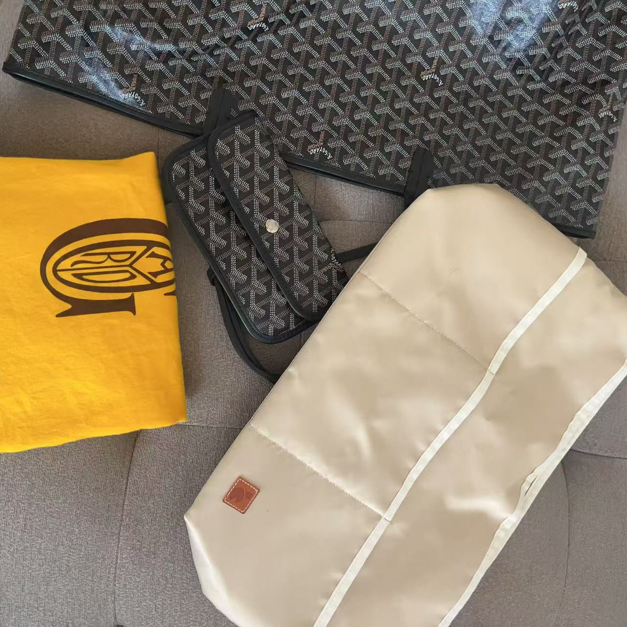 A luxury Goyard bag set arranged on a gray fabric surface, including a large black and gray patterned tote bag, a matching small bag, a folded bright yellow Goyard dust bag, and a cream-colored fabric pouch.