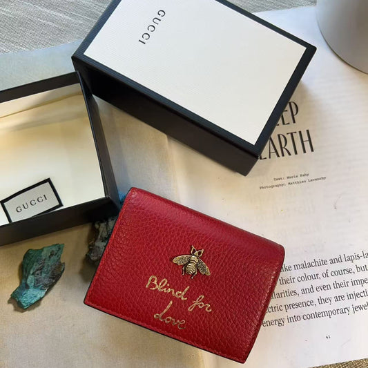 A red textured leather Gucci wallet with gold embroidery reading 'Blind for love' and a gold bee motif, displayed with its black and white Gucci box and an open Gucci box on a beige surface.