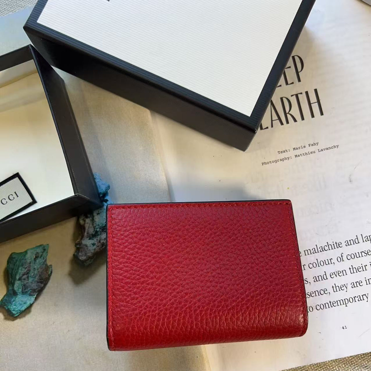 A red pebbled leather bifold wallet is displayed on an open book, with its black and white Gucci packaging boxes and a small turquoise stone nearby.