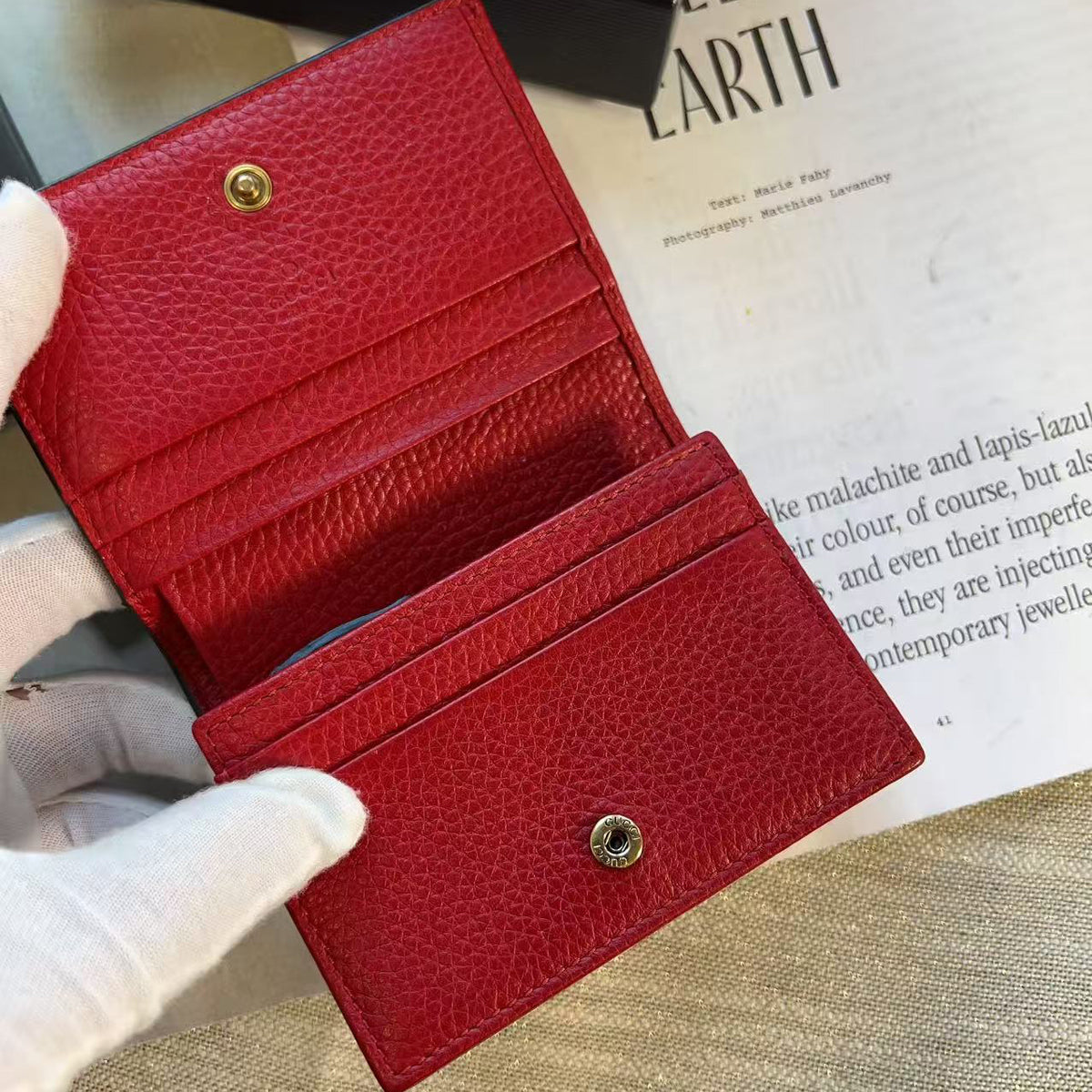 A red textured leather card holder wallet held open by a hand wearing a white glove, with a small gold-tone snap button closure. The wallet is shown against a styled background featuring a magazine page and a black box corner.
