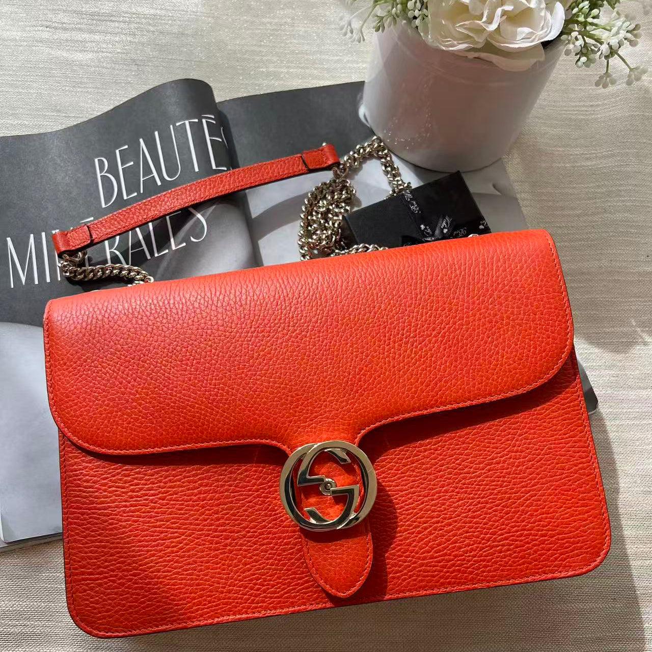 A bright orange-red textured leather handbag with a gold-tone interlocking G logo clasp, placed on top of a magazine with a floral pot in the background.