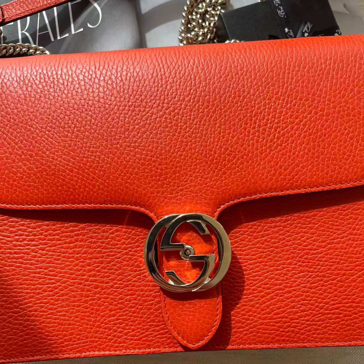 Close-up of a bright orange-red pebbled leather handbag with a gold-tone interlocking G logo clasp, showing part of the chain strap and a branded dust bag.