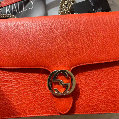 Close-up of a bright orange-red pebbled leather handbag with a gold-tone interlocking G logo clasp, showing part of the chain strap and a branded dust bag.