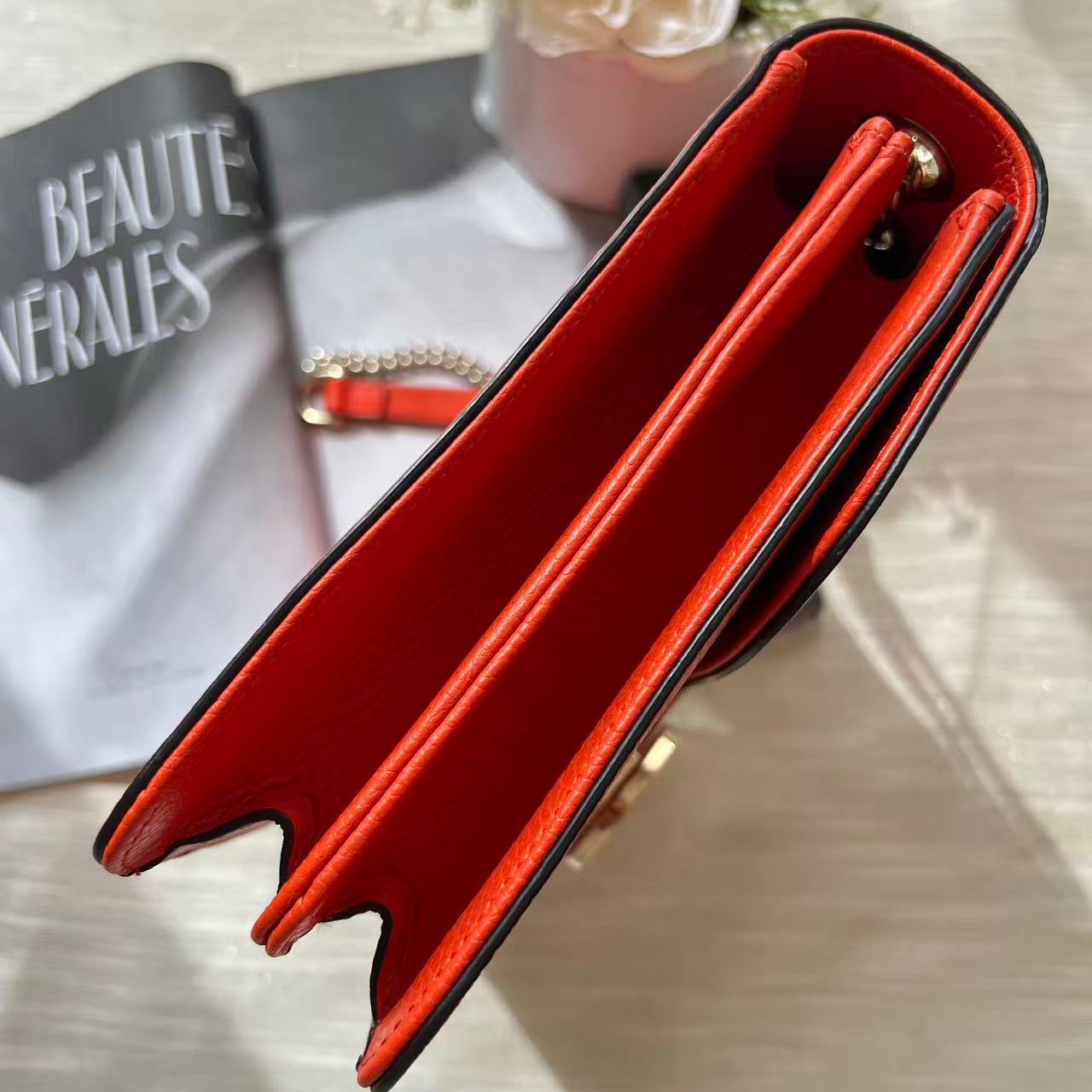A close-up of a red leather clutch or wallet-style handbag shown open, with black trim and a small gold-tone hardware detail. The bag is photographed on a light fabric surface with a magazine and flowers in the background.