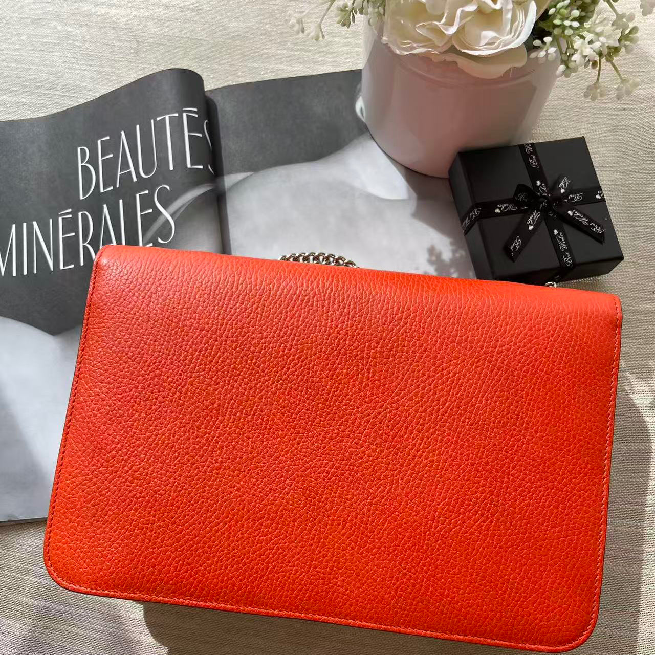 A bright orange-red textured leather clutch bag with a silver chain strap, styled on a light gray fabric surface with a magazine, a small black gift box, and a pink vase with flowers.