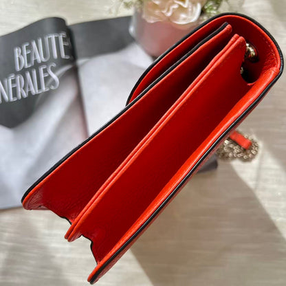 A close-up of a red leather wallet or card holder with black trim, photographed on a light fabric surface with a magazine and flowers in the background.