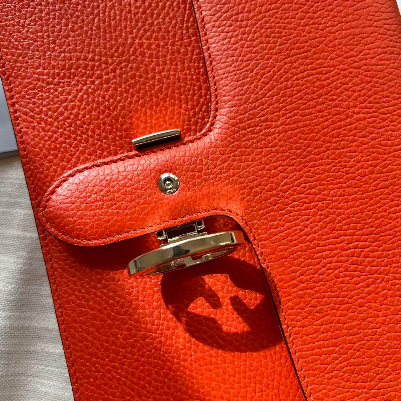 Close-up of a bright orange-red pebbled leather clutch or wallet with gold-tone hardware and a turn-lock closure, photographed on a light neutral surface.