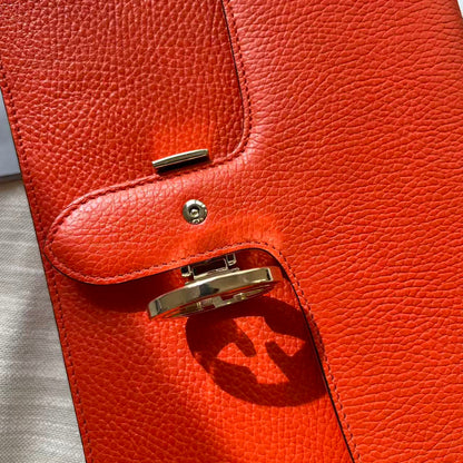 Close-up of a bright orange-red pebbled leather clutch or wallet with gold-tone hardware and a turn-lock closure, photographed on a light neutral surface.