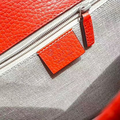 Close-up of a Gucci leather and canvas zippered bag showing the orange-red pebbled leather zipper pull tab with the embossed Gucci logo and 'MADE IN ITALY' text on a gray textured canvas background.