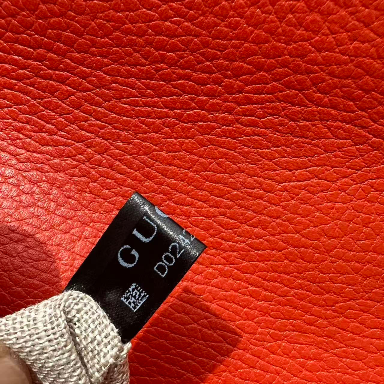 Close-up of a textured bright red leather item with a small black fabric tag attached, held by a finger. The tag shows partial white text including 'GU' and 'D225'.