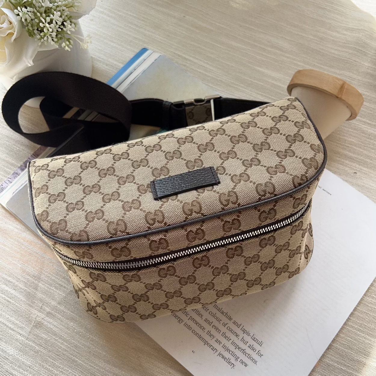 A beige and brown monogram canvas crossbody bag with a black strap, resting on a light wood surface over a white sheet of paper.