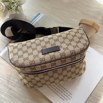 A beige and brown monogram canvas crossbody bag with a black strap, resting on a light wood surface over a white sheet of paper.