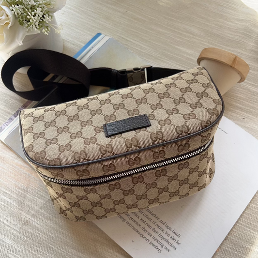 A beige and brown monogram canvas crossbody bag with a black strap, resting on a light wood surface over a white sheet of paper.
