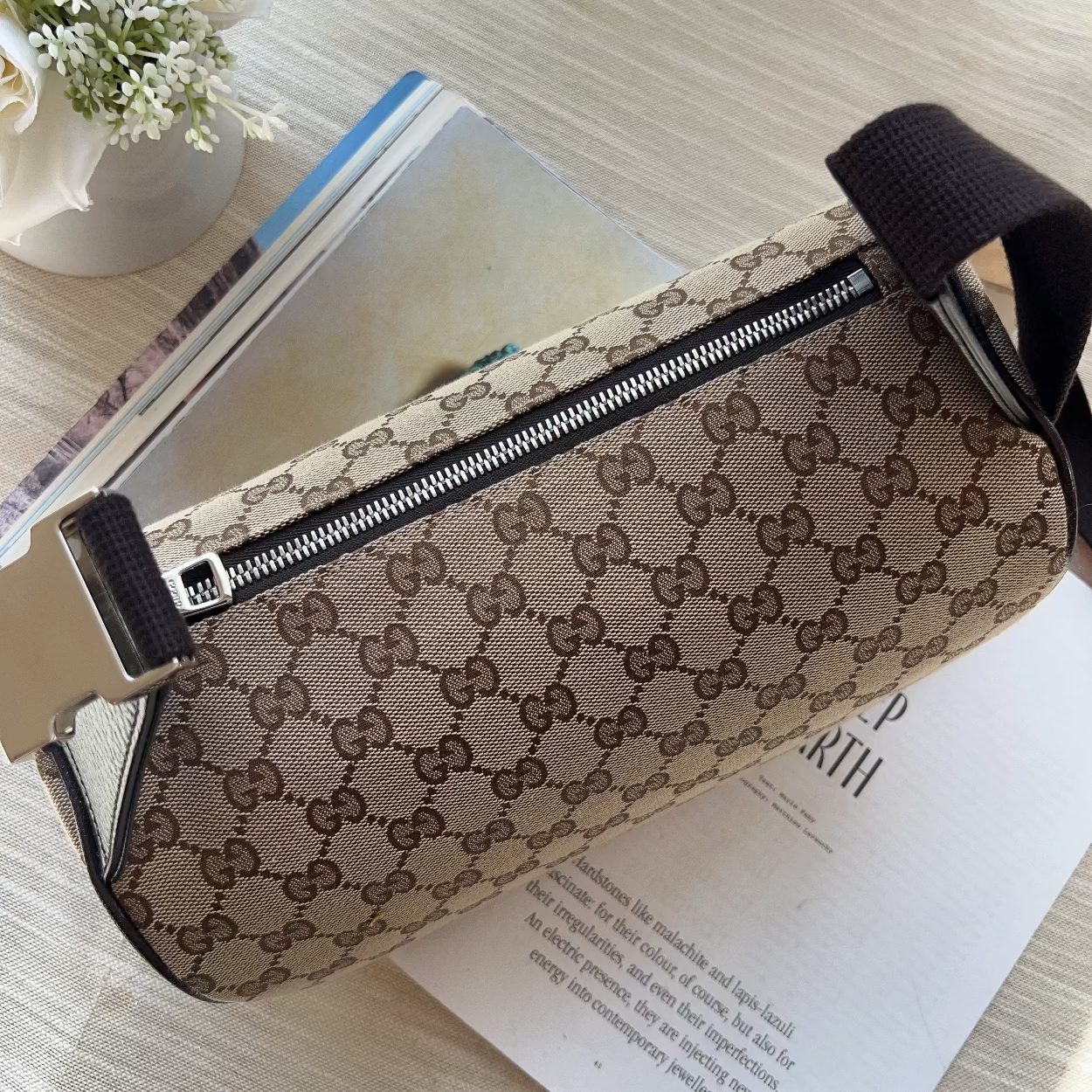 A Gucci monogram canvas cylindrical pouch with a black zipper and black strap, resting on a light wood surface over an open book.