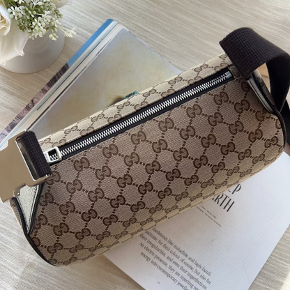 A Gucci monogram canvas cylindrical pouch with a black zipper and black strap, resting on a light wood surface over an open book.