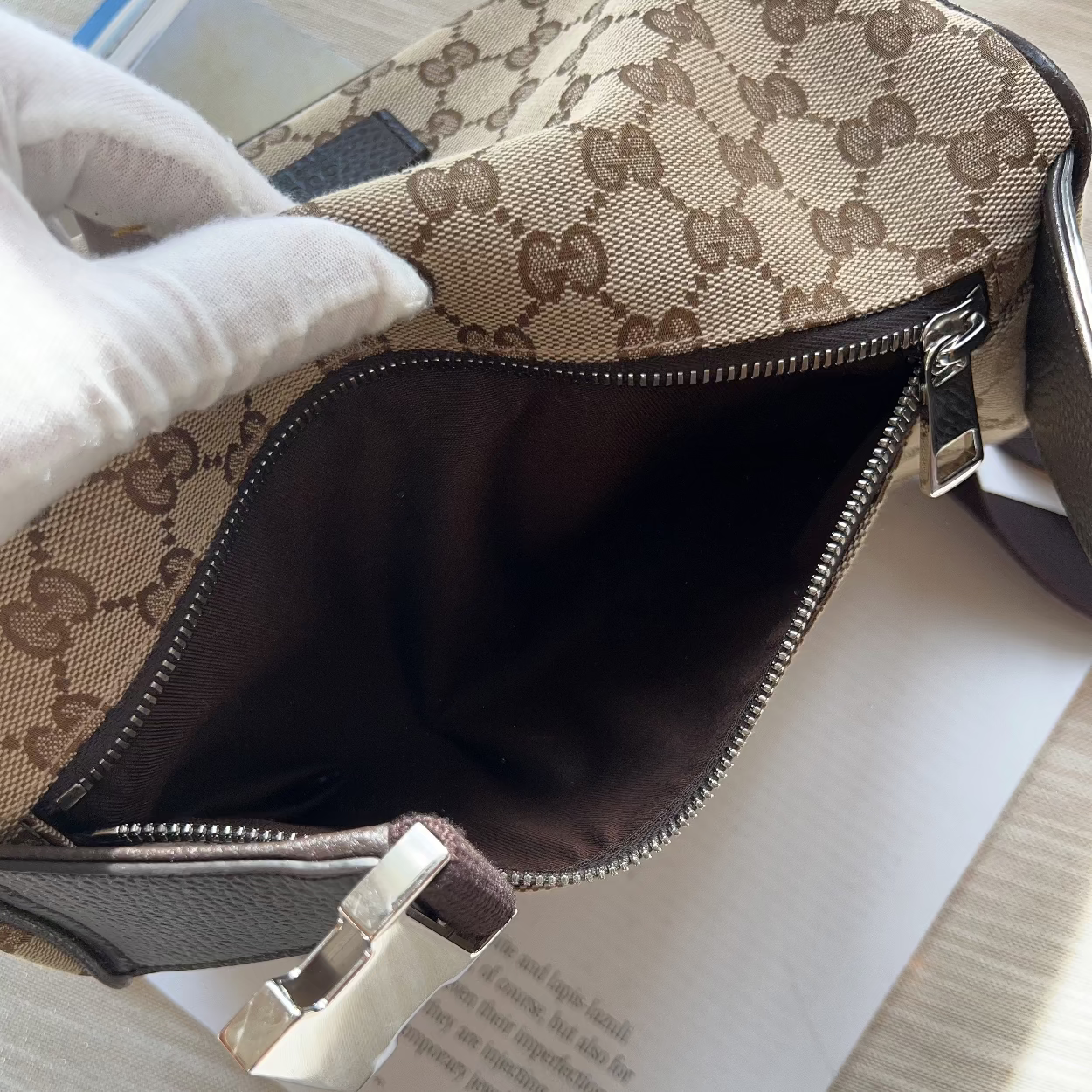 Close-up of a beige and brown monogram handbag with a white-gloved hand holding the open top, showing the dark brown interior and silver zipper hardware on a light wood surface.