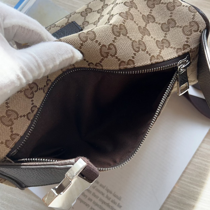 Close-up of a beige and brown monogram handbag with a white-gloved hand holding the open top, showing the dark brown interior and silver zipper hardware on a light wood surface.