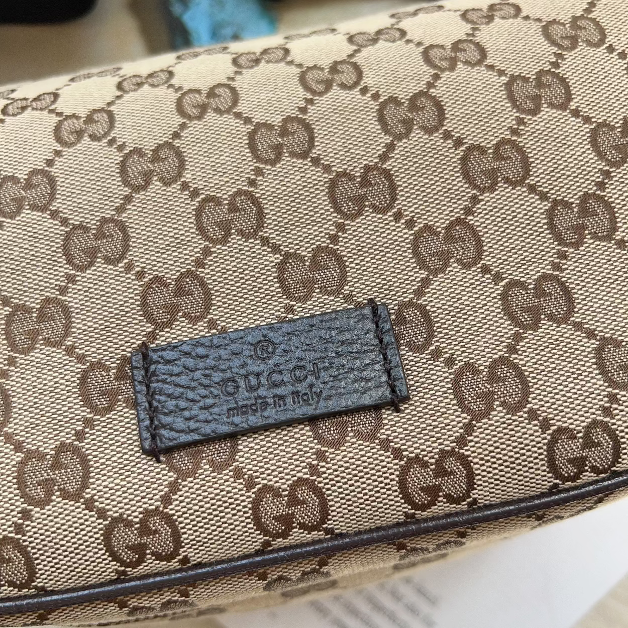 Close-up of a Gucci monogram bag in beige and brown canvas with a dark brown leather brand patch reading 'GUCCI MADE IN ITALY'.