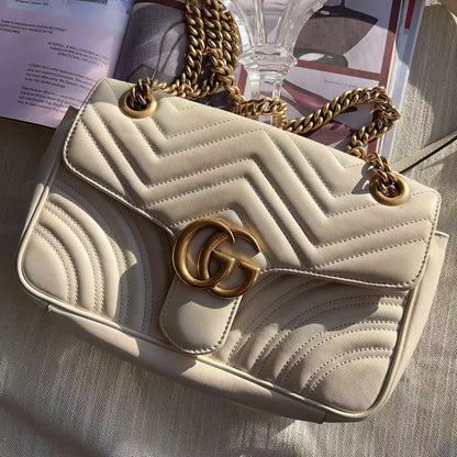 Beige handbag with gold chain strap and clasp on a textured surface.