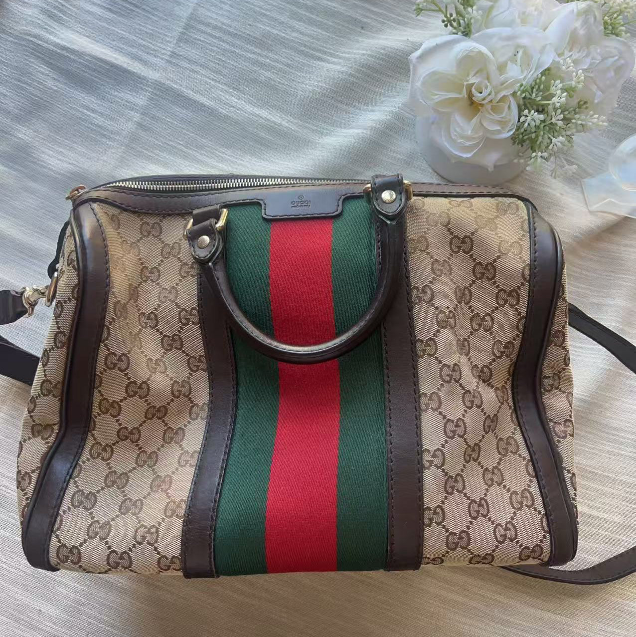 A Gucci Saba handbag in the classic GG Supreme canvas, featuring the iconic green and red web stripe down the center, dark brown leather trim, and top handle, displayed on a light-colored wooden surface next to a vase of white flowers.