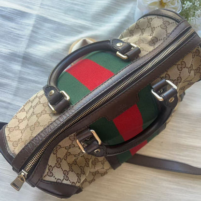 A Gucci dome-shaped shoulder bag in the classic GG Supreme canvas, featuring dark brown leather trim, gold-tone hardware, and the signature green and red web stripe pattern on the front, displayed on a light-colored fabric surface.