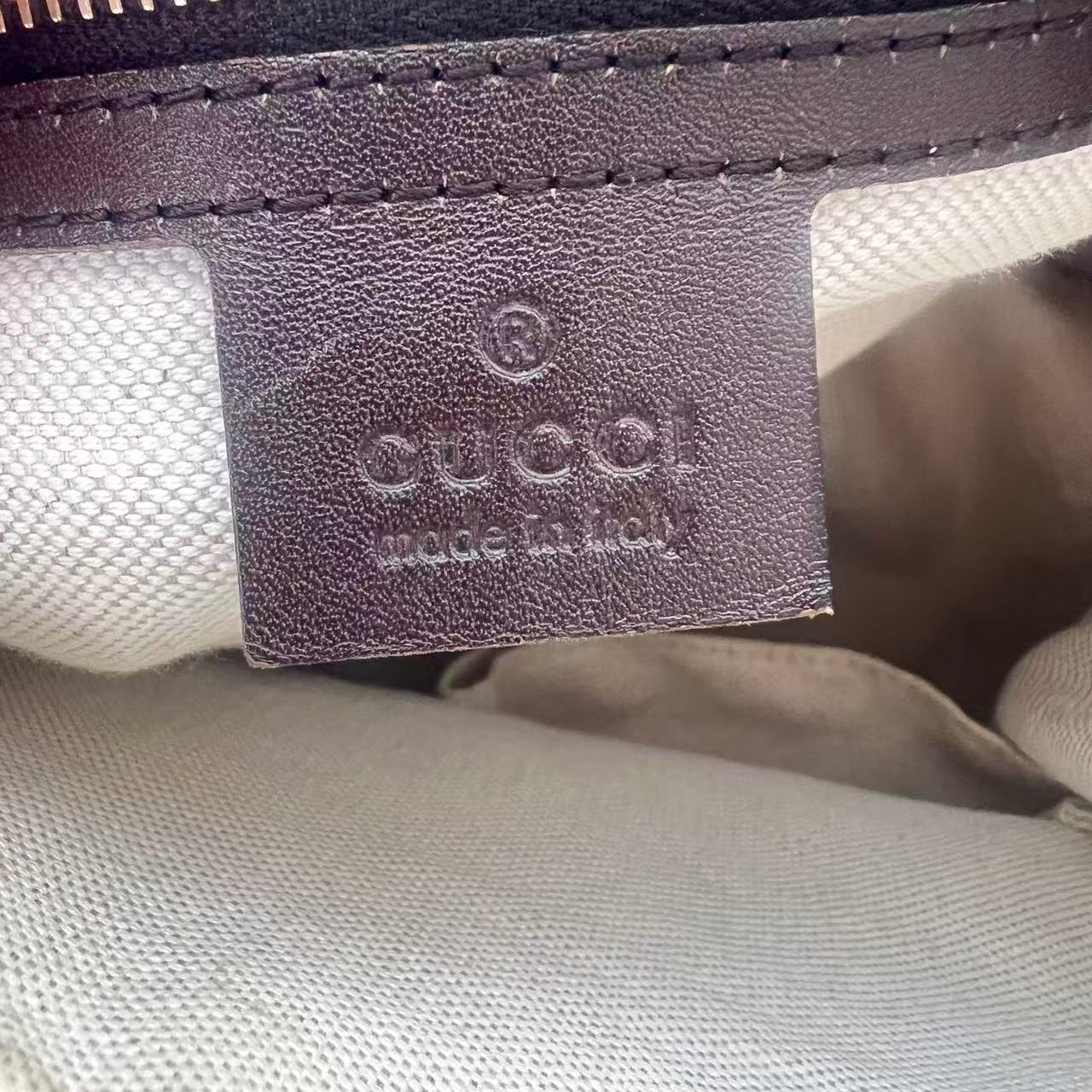 Close-up of a Gucci leather interior label with 'GUCCI made in Italy' embossed on a brown leather tab, showing part of a beige canvas body and a gold zipper.