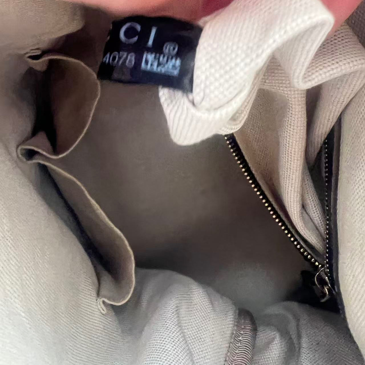 Close-up of a hand holding a black fabric tag sewn onto the inside of a light beige canvas bag, with a zipper and interior lining visible.