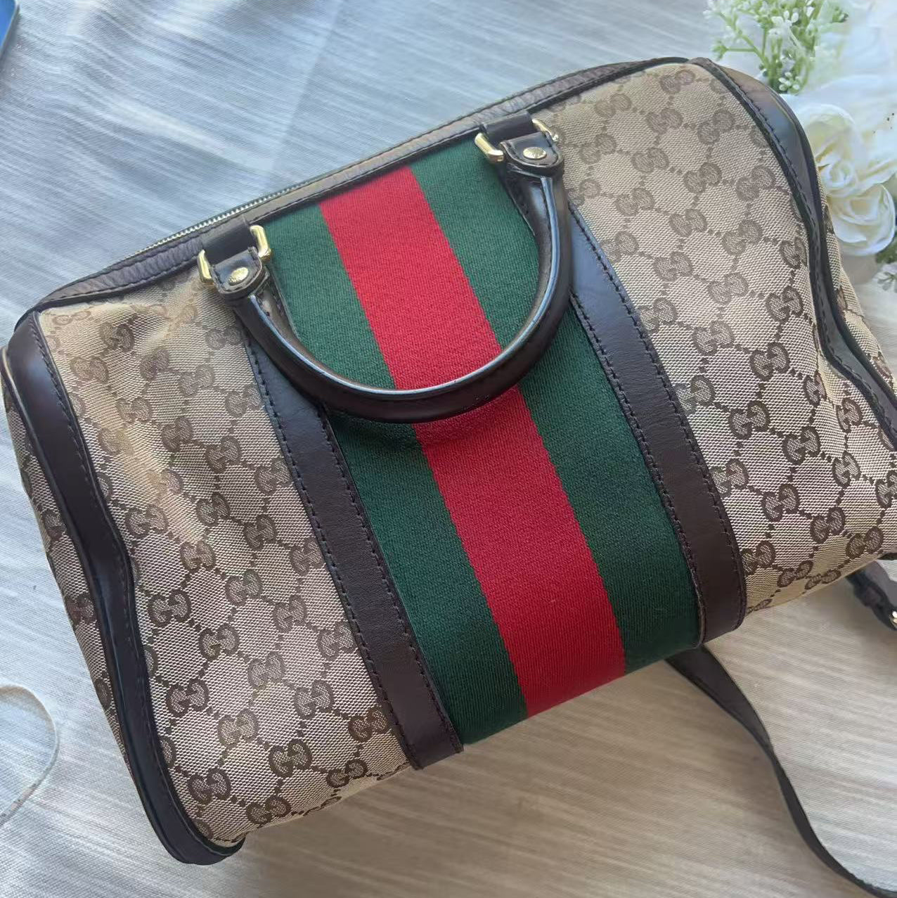 A Gucci monogram canvas duffel bag with the signature green and red web stripe down the center, dark brown leather trim, and a top handle, photographed on a light fabric surface.