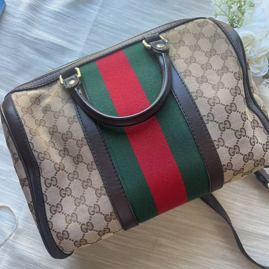 A Gucci monogram canvas duffel bag with the signature green and red web stripe down the center, dark brown leather trim, and a top handle, photographed on a light fabric surface.