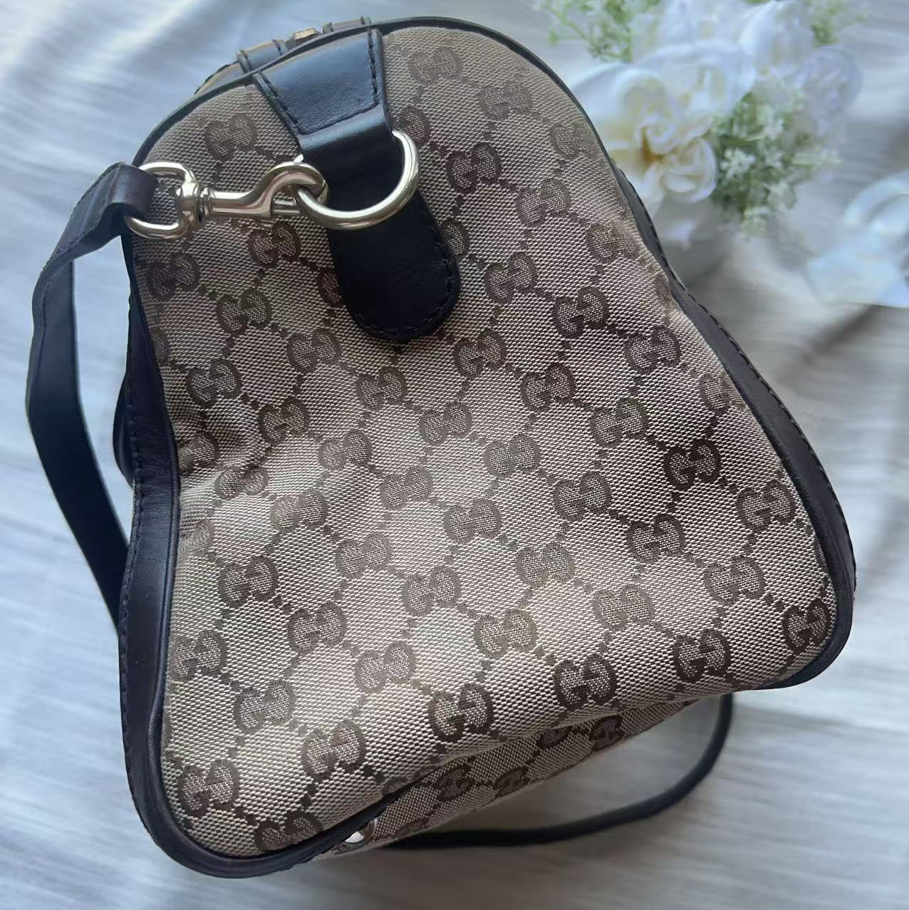 A Gucci shoulder bag in beige and brown GG Supreme canvas with black leather trim and a gold-tone ring detail, resting on a light gray fabric surface with white flowers in the background.