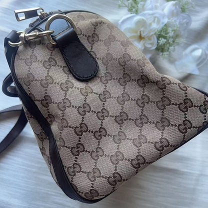 A Gucci shoulder bag in beige and brown GG Supreme canvas with black leather trim and gold-tone hardware, photographed on a light fabric surface with white flowers in the background.