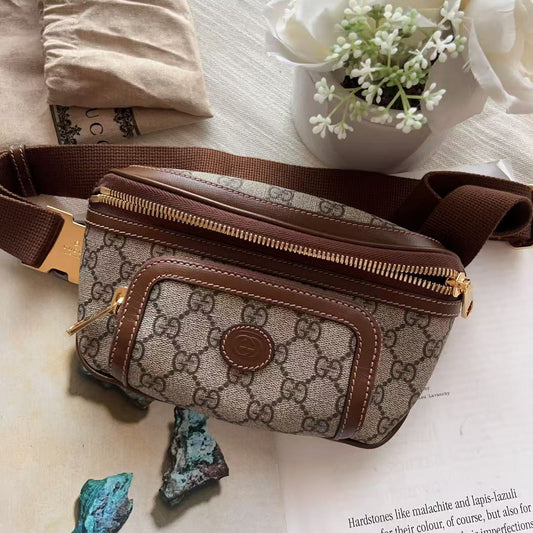 A Gucci monogram canvas fanny pack with brown leather trim and a matching brown strap, styled on a light surface with a beige pouch, white flowers, and small blue-green stones.