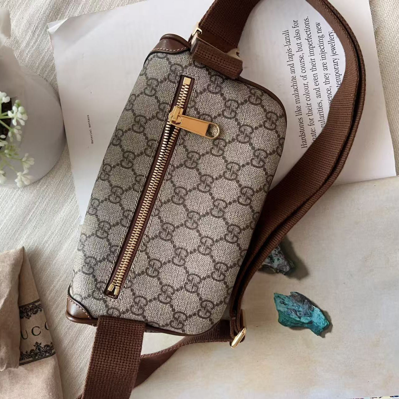 A Gucci monogram canvas shoulder bag with brown leather trim and a gold-tone zipper, styled on a textured cream fabric with a scarf, white rose, and printed paper.
