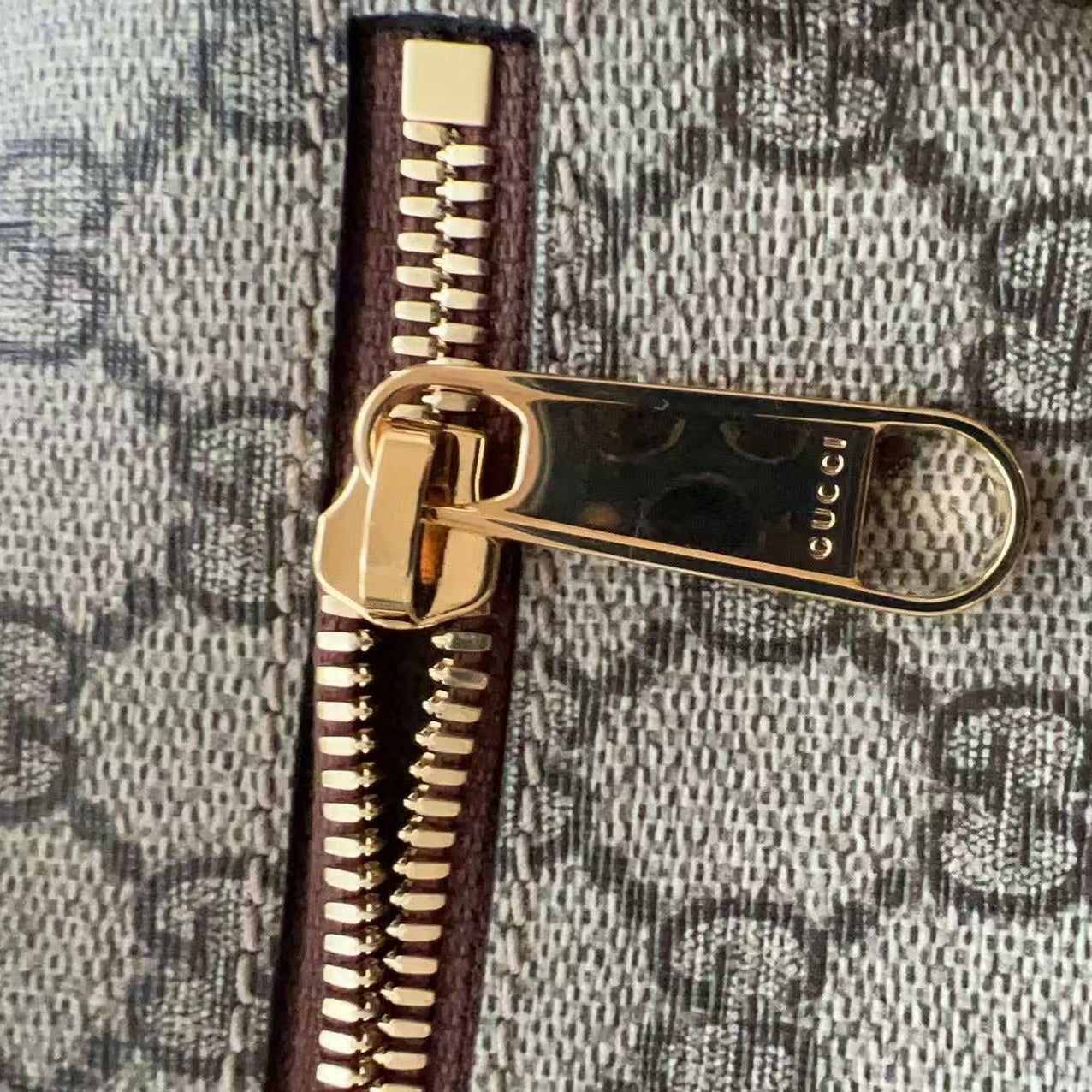 Close-up of a Gucci zipper pull and monogram canvas detail, showing the gold-tone zipper pull engraved with 'GUCCI' and the repeating GG monogram pattern.