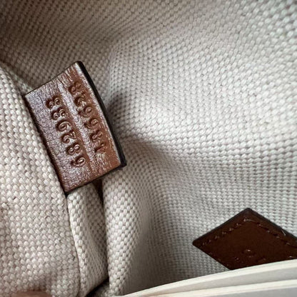 Close-up of a textured beige canvas or woven fabric interior with a brown leather tag showing embossed numbers, and part of another brown leather detail in the lower right corner.